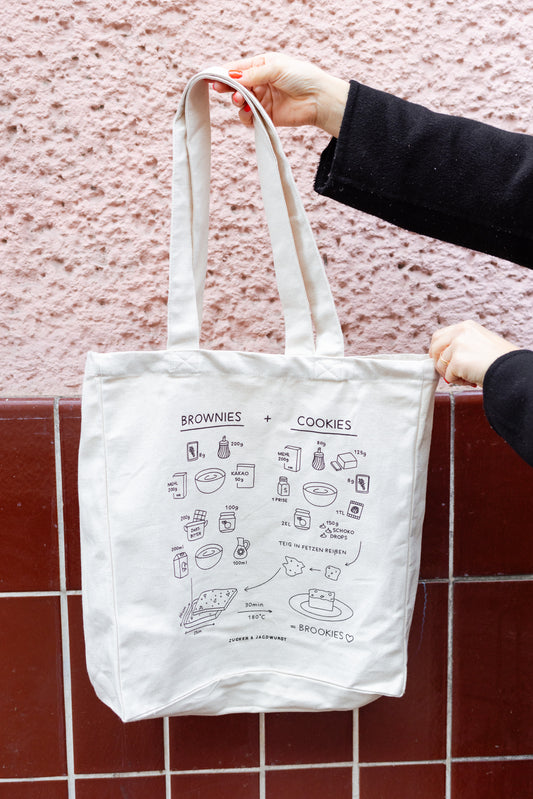 Weißer Jutebeutel mit Rezept ‚Brookies‘, gehalten von einer Hand, vor rosa und dunkelrotem Hintergrund