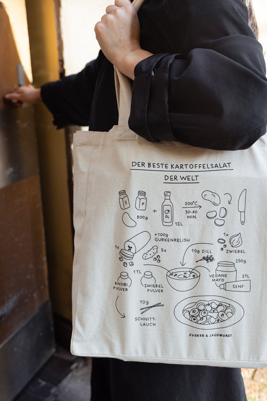Person trägt weißen Jutebeutel mit Rezept ‚Der beste Kartoffelsalat der Welt‘, schwarze Jacke, draußen vor der Haustür