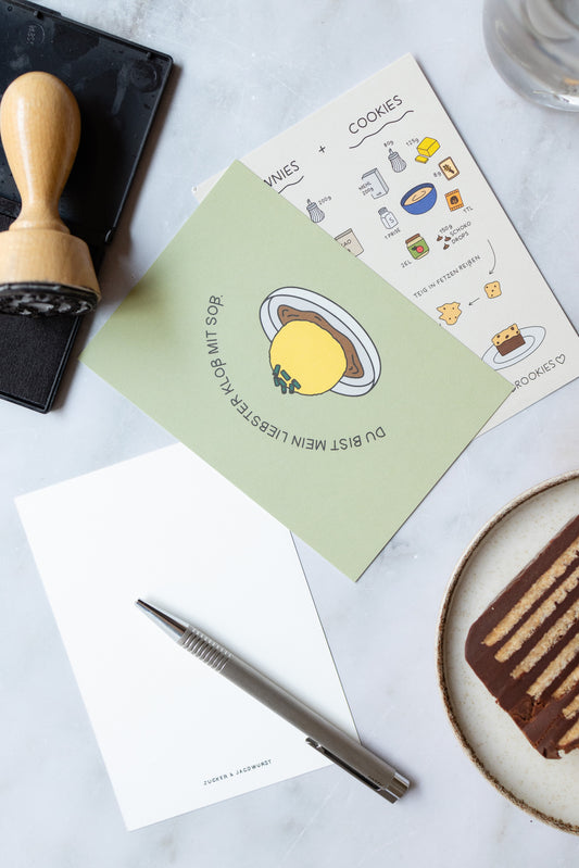 Postkarten auf einem Tisch: vorne ‚Du bist mein liebster Kloß mit Soß‘, dahinter ‚Brookies‘, unten ein Stück Kalter Hund Kuchen, links eine leere Postkarte mit Stift, rechts ein Stempel mit Stempelset