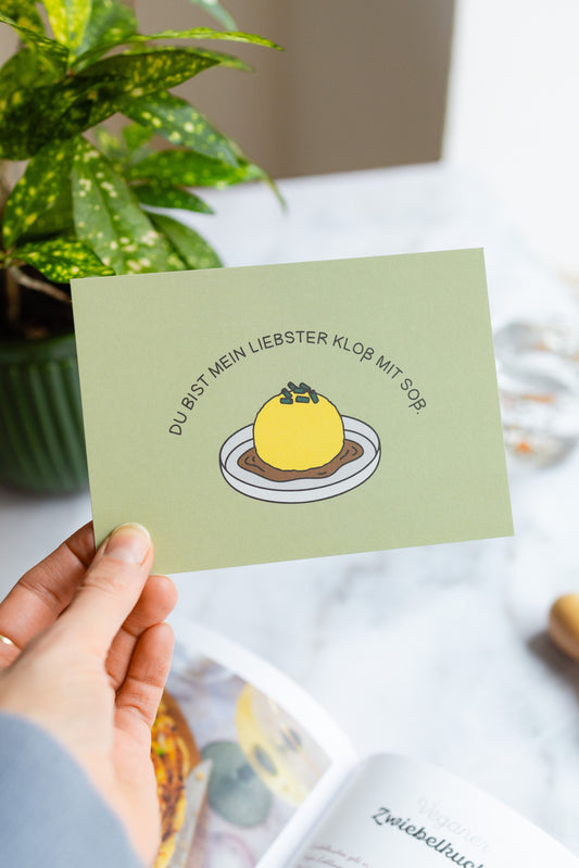 Hand hält eine ‚Du bist mein liebster Kloß mit Soß‘ Postkarte, im Hintergrund links eine Pflanze und rechts ein Wasserglas