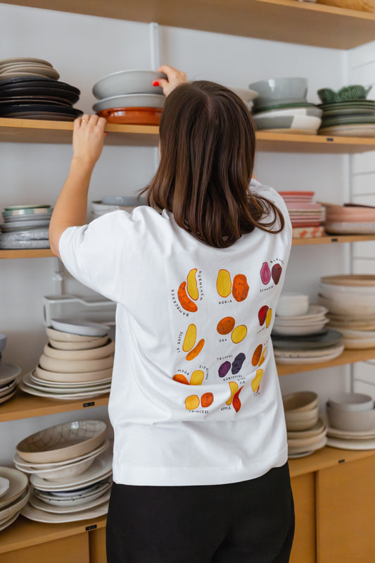 Rückenansicht des weißes T-Shirt mit Illustration ‚Kartoffeln‘ getragen von einer Person, die gerade Teller verräumt