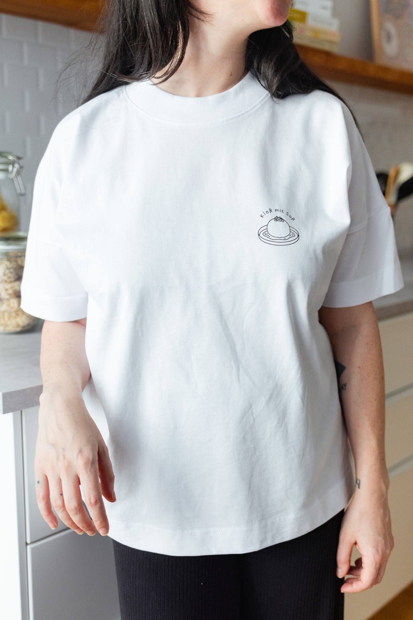 Person trägt weißes T-Shirt mit Illustration ‚Kloß mit Soß‘ und ist gerade in der Küche