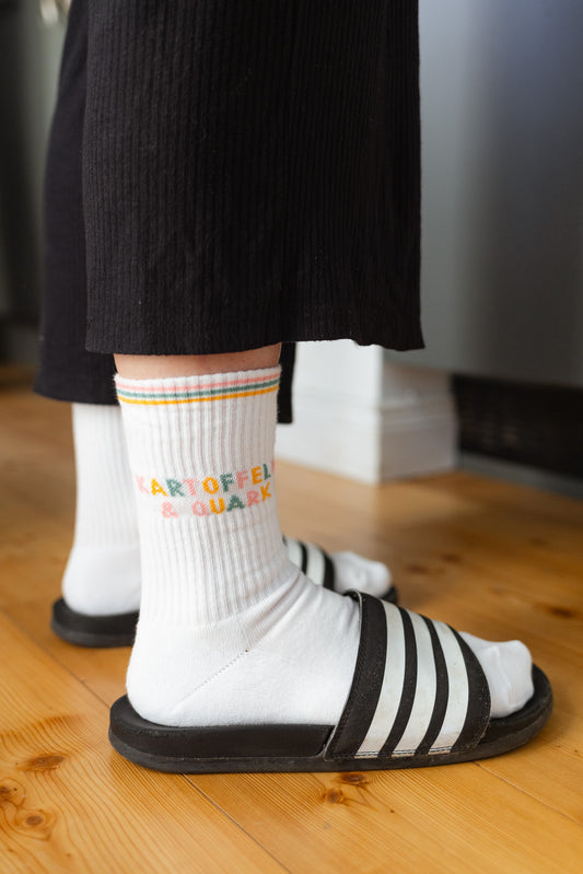 Weiße Tennissocken mit buntem Schriftzug ‚Kartoffeln & Quark‘, getragen von einer Person mit schwarz-weißen Badelatschen