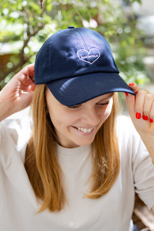 Vordere Ansicht einer dunkelblauen Kappe mit weißer Herz-Stickerei ‚Kartoffeln‘, getragen von einer Person