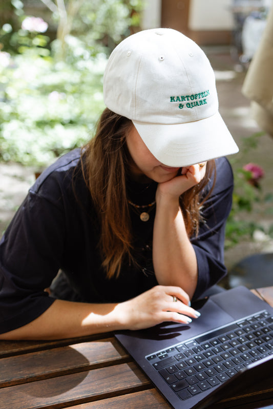 Vordere Ansicht einer weißen Kappe mit dunkelgrüner Stickerei ‚Kartoffeln & Quark‘, getragen von einer Person