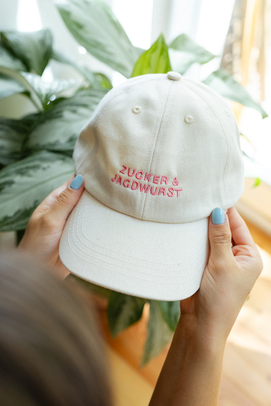Vordere Ansicht einer weißen Kappe mit pinker Stickerei ‚Zucker & Jagdwurst‘, in der Hand