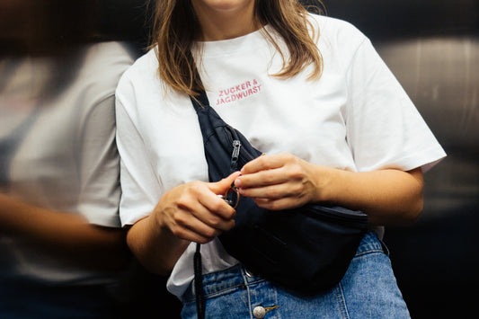 Person trägt weißes T-Shirt ‚Zucker & Jagdwurst‘, schwarze Bauchtasche und Jeans, steht im Aufzug