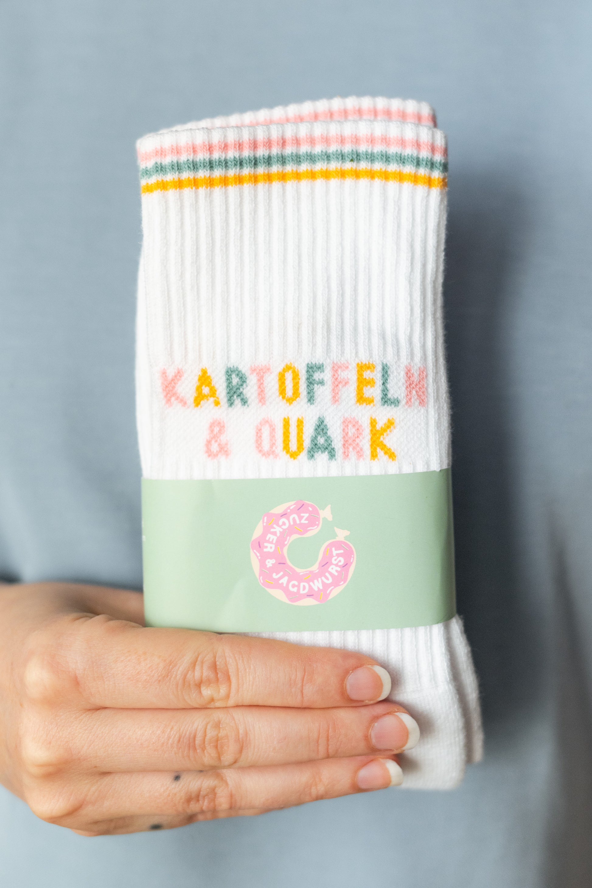 Hand hält weiße Tennissocken mit buntem Schriftzug ‚Kartoffeln & Quark‘ in der Verpackung