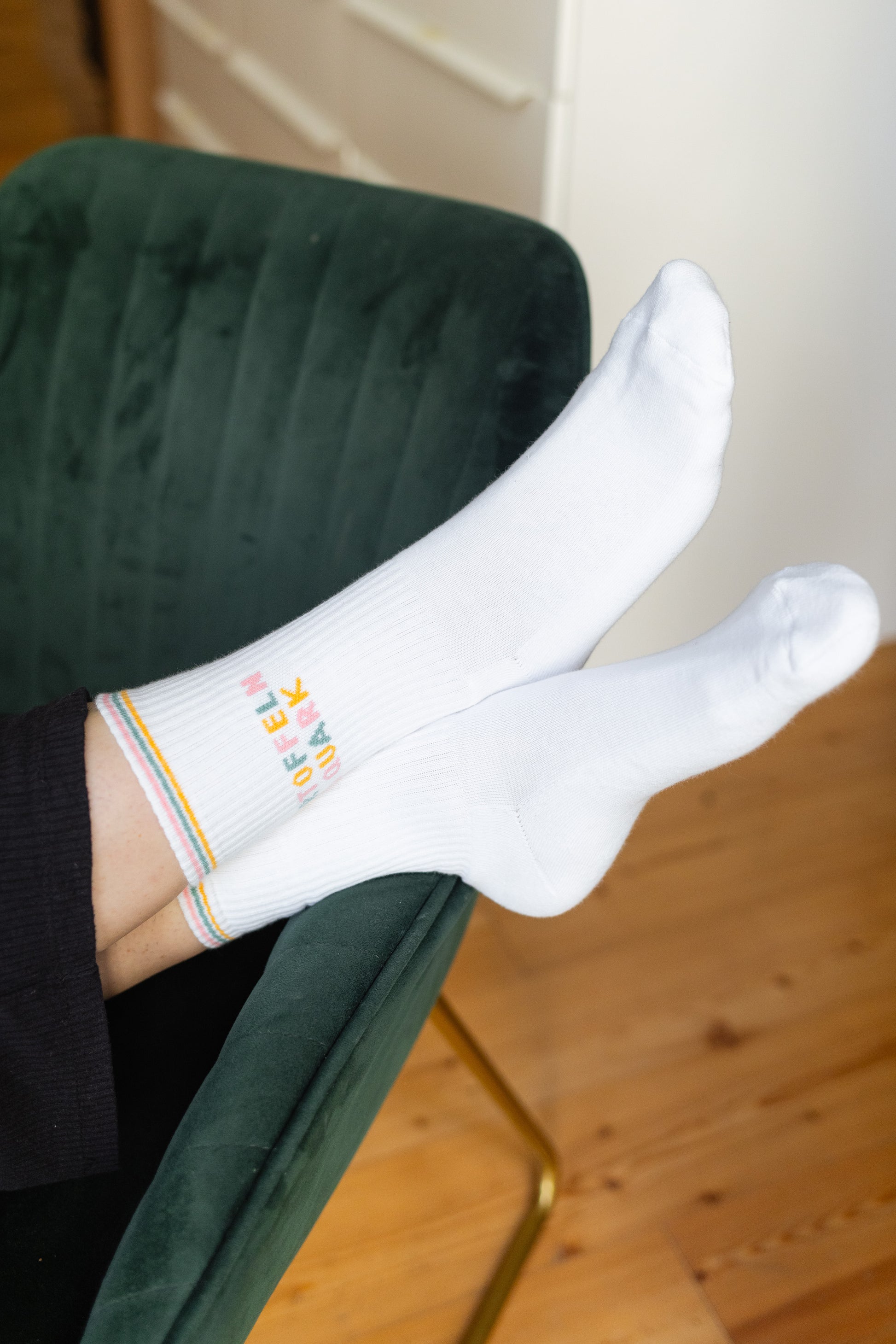 Weiße Tennissocken mit buntem Schriftzug ‚Kartoffeln & Quark‘, getragen von einer Person, vor grüner Samt-Couch