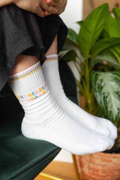 Weiße Tennissocken mit buntem Schriftzug ‚Kartoffeln & Quark‘, getragen von einer Person, vor grüner Samt-Couch