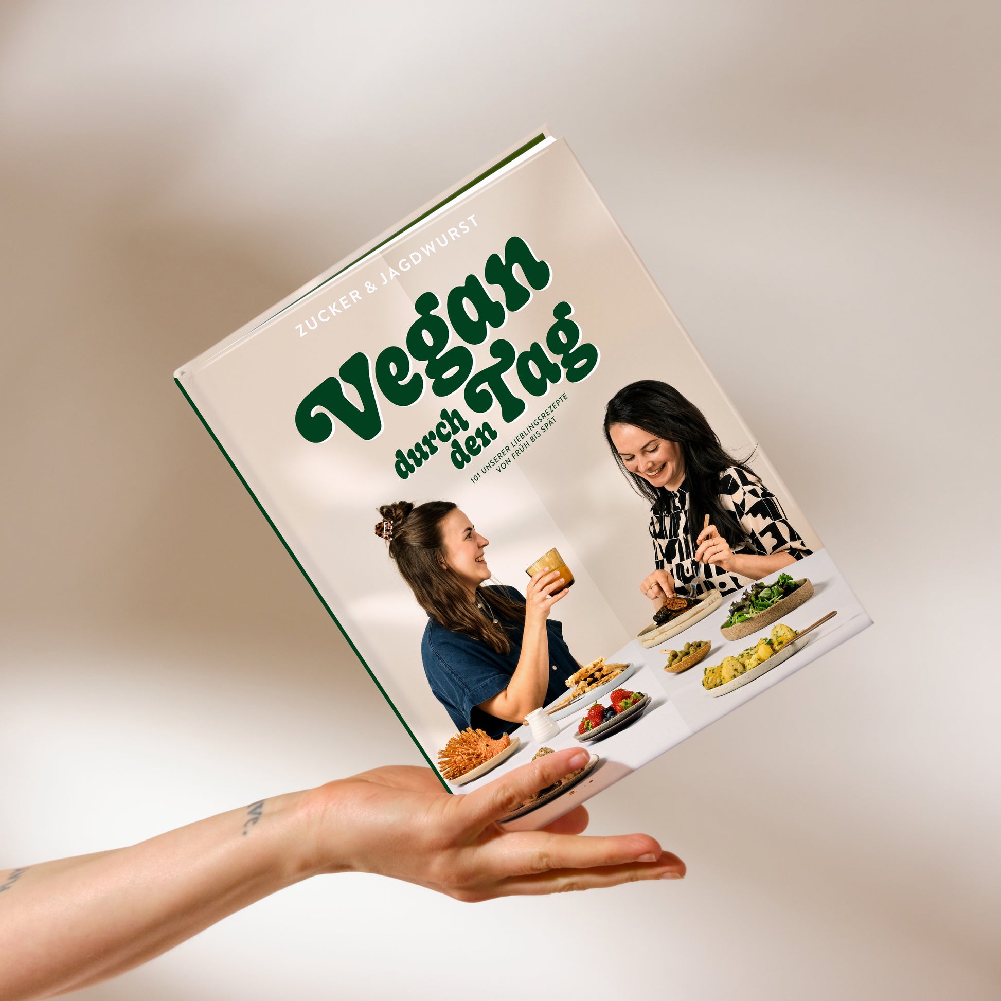 Kochbuch ‚Vegan durch den Tag‘ auf beigem Hintergrund mit Hand