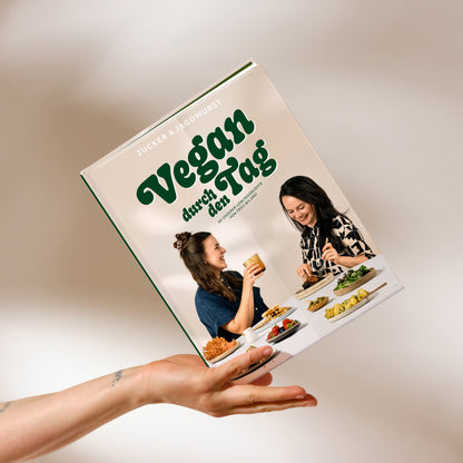 Kochbuch ‚Vegan durch den Tag‘ auf beigem Hintergrund mit Hand