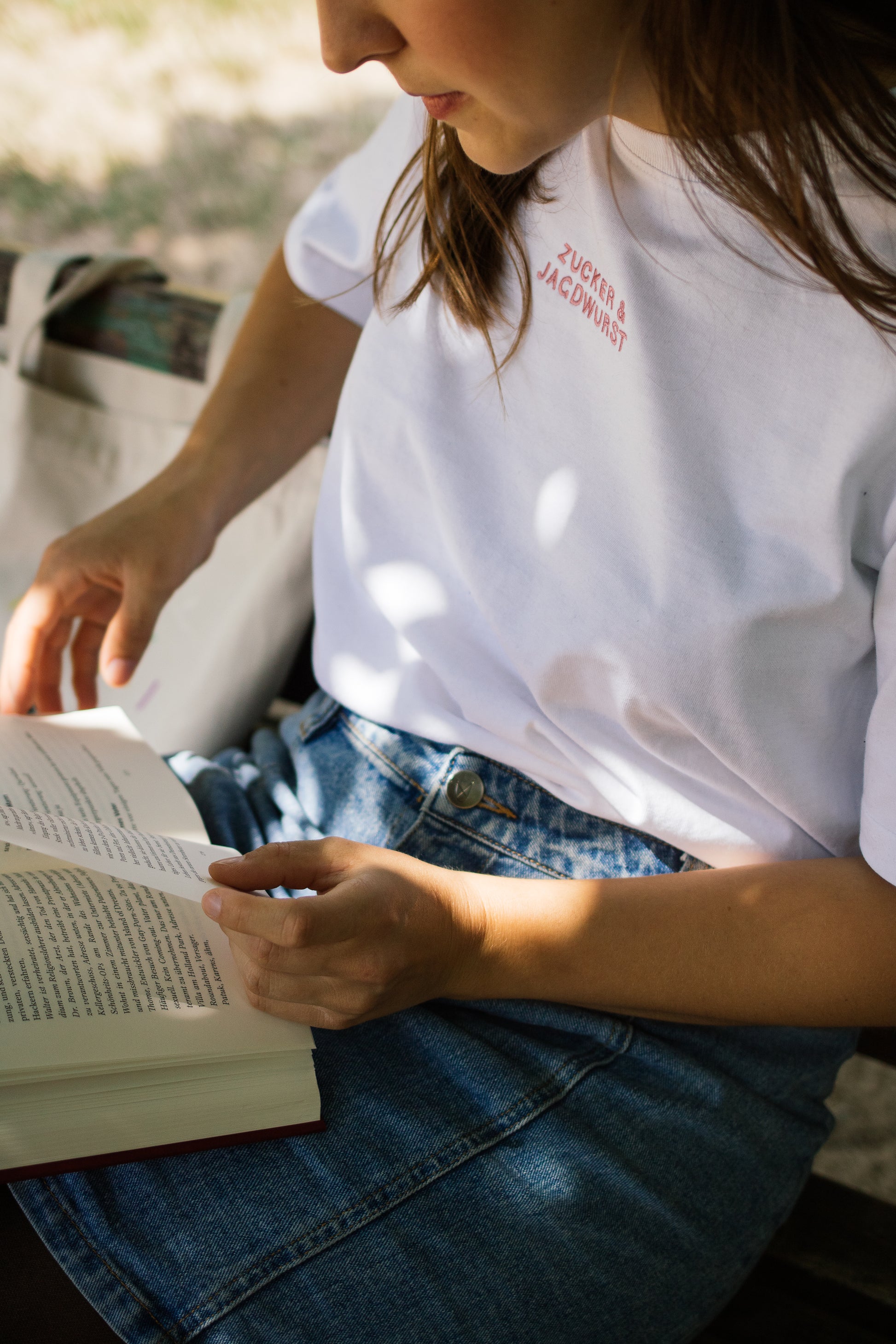 Person trägt weißes T-Shirt ‚Zucker & Jagdwurst‘, Jeans-Rock, sitzt auf einer Bank und liest ein Buch
